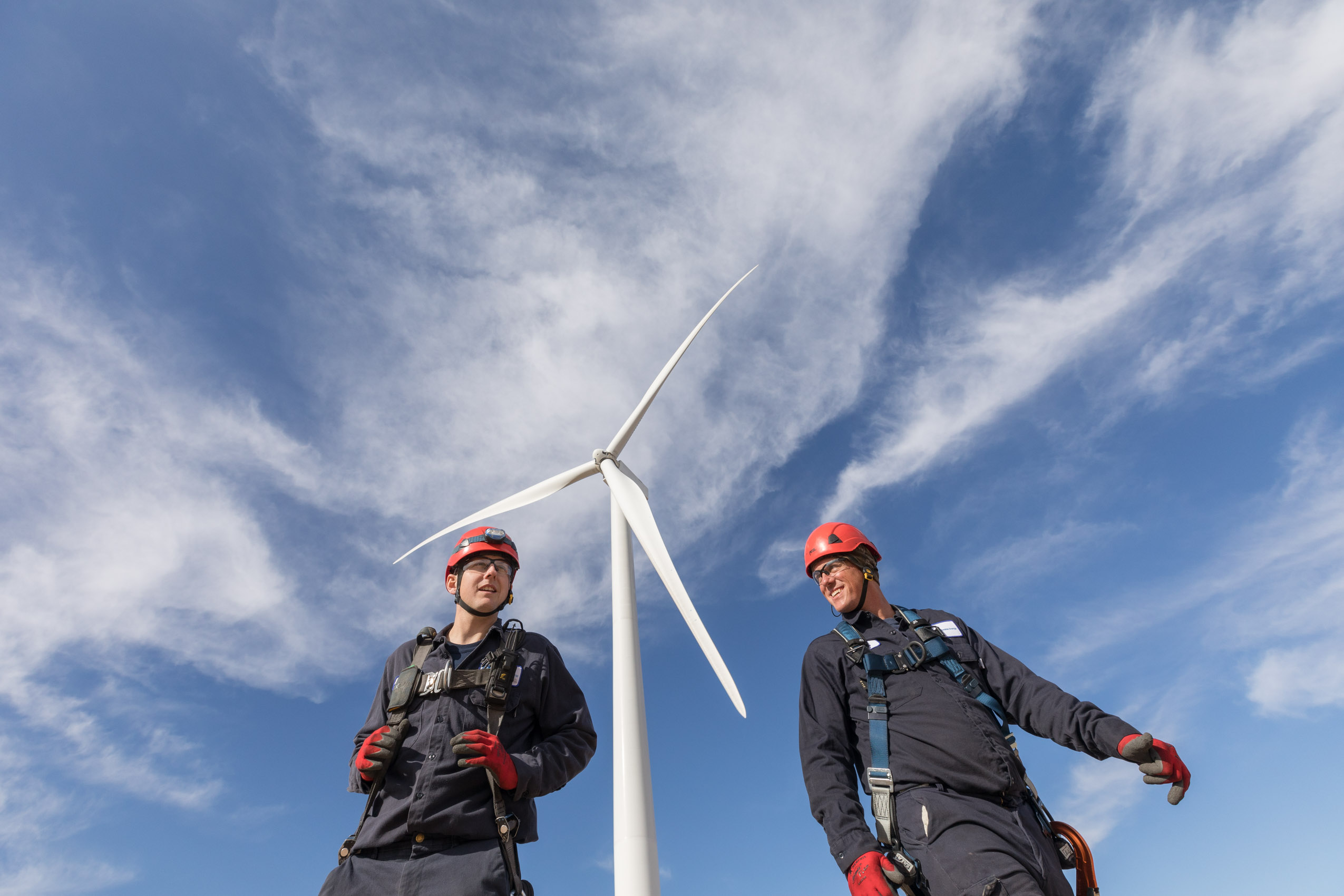 Two Amazon Wind techs walking below wind turbine smiling with safety gear on.